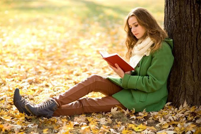 Una donna legge un libro appoggiata a un albero, tra le foglie autunnali.